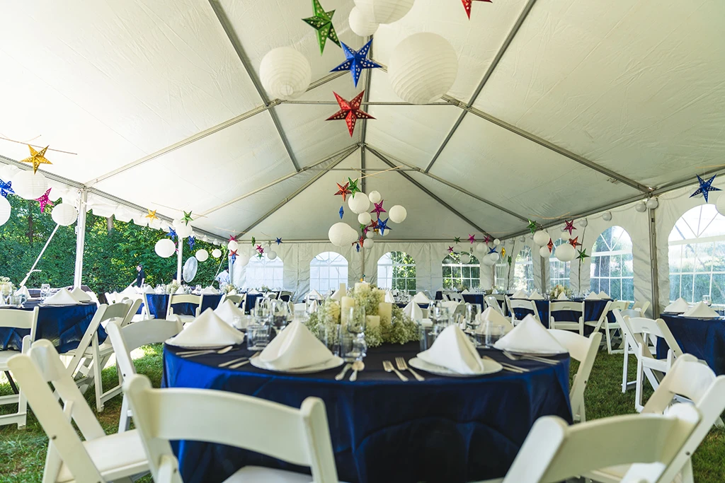 Tent reception setup with blue linens, white chairs, and colorful hanging lanterns