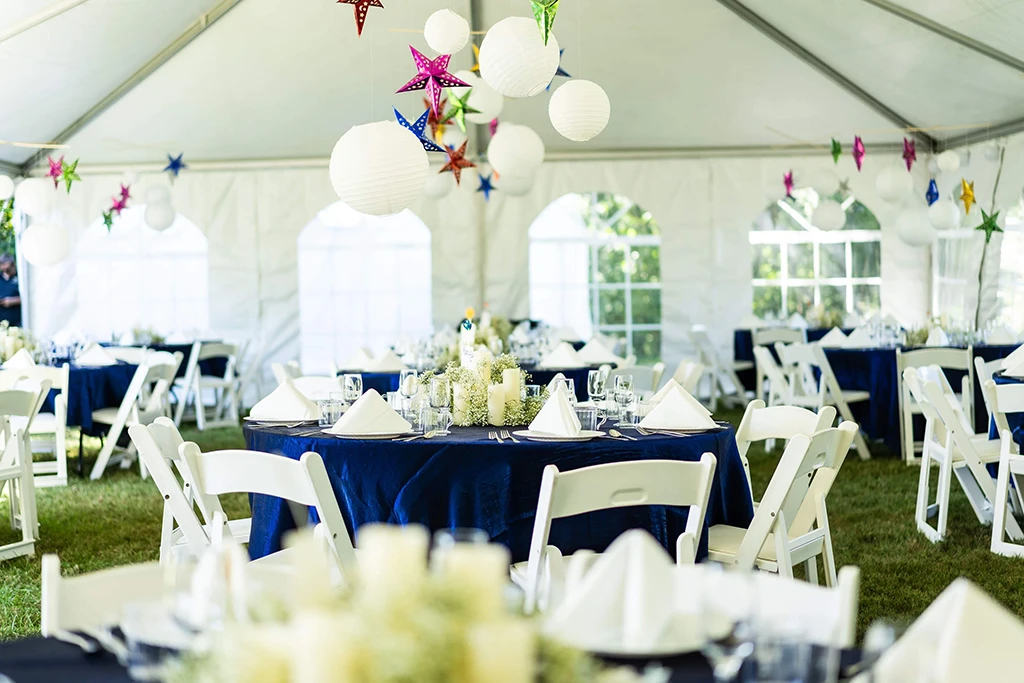 Tent reception view with white chairs, round tables, and colorful lanterns