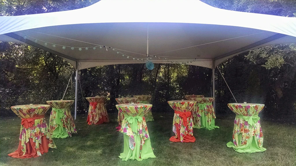 Frame tent over high-top tables with colorful floral covers in a wooded backyard